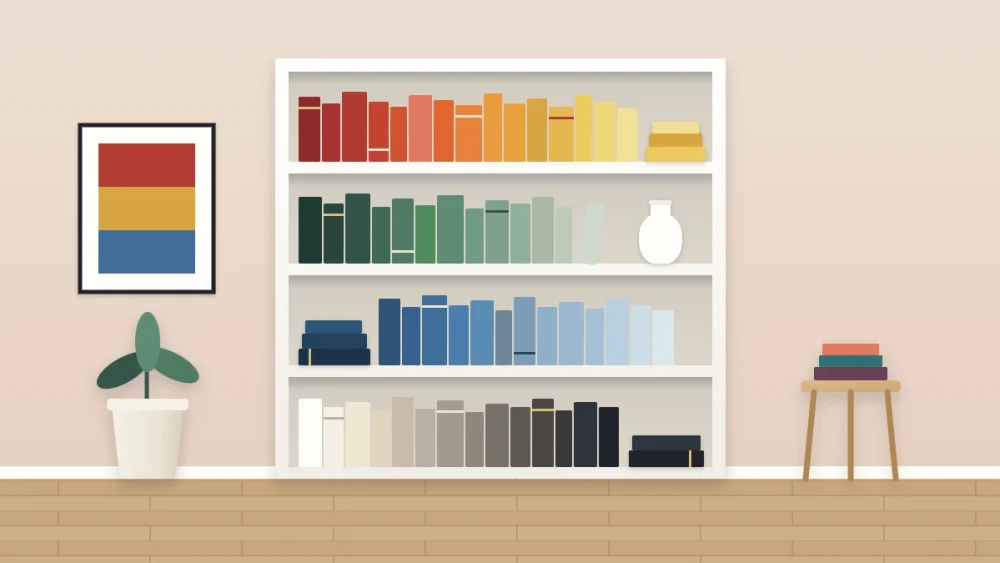 A white bookcase with four shelves of plain-spined books sorted by color, from reds and yellows to greens, blues and neutrals, with a small white vase